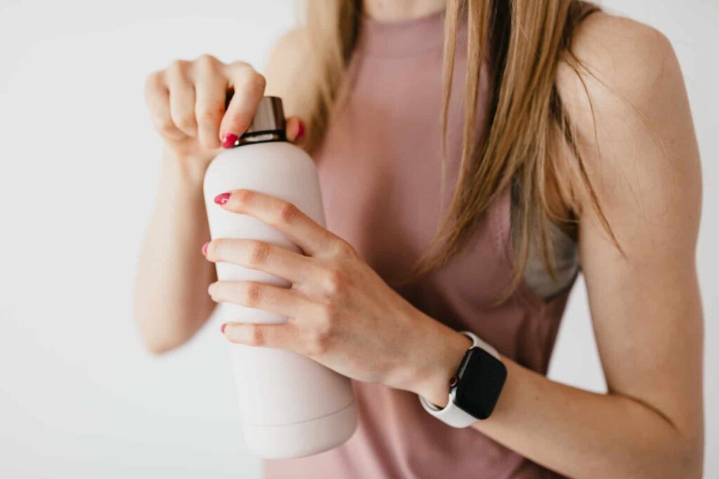 Crop faceless young female in casual outfit wearing smart watch opening cosmetic bottle on white background